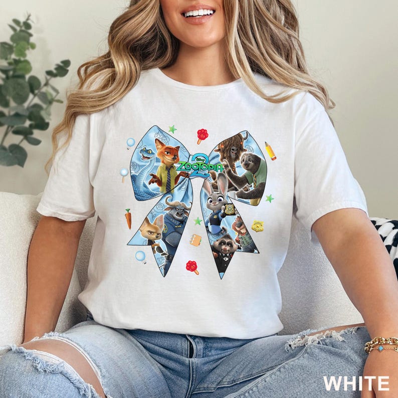 Op de afbeelding: Wit T-shirt met een groot blauw strikdesign met personages uit de animatiefilm "Zootropolis". De strik bevat afbeeldingen van Nick Wilde, Judy Hopps en andere personages. Het woord "Zootropolis" staat in groene tekst. Het woord "WHITE" staat onderaan.
