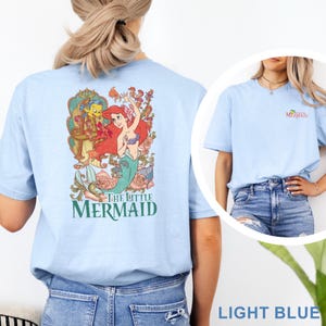 May include: A light blue t-shirt featuring a large graphic on the back of Ariel and Flounder from The Little Mermaid surrounded by floral elements and the text The Little Mermaid. The front chest area displays a small logo with the same text.