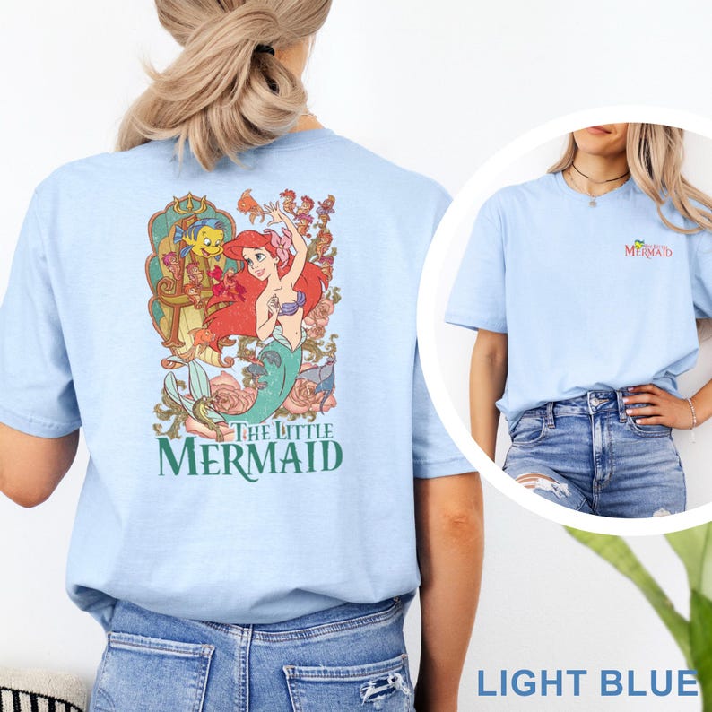 May include: A light blue t-shirt featuring a large graphic on the back of Ariel and Flounder from The Little Mermaid surrounded by floral elements and the text The Little Mermaid. The front chest area displays a small logo with the same text.