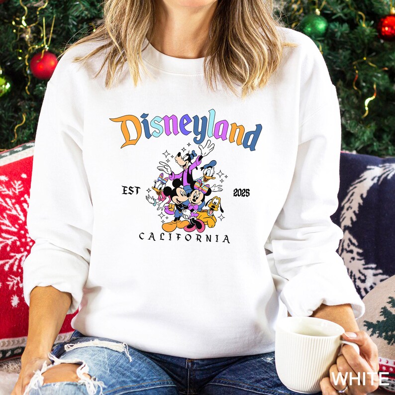 Puede incluir: Sudadera blanca con el logotipo de Disneyland en naranja y azul, y un gr&aacute;fico de personajes de Disney. El texto "EST 2025 CALIFORNIA" tambi&eacute;n est&aacute; impreso. La sudadera es de cuello redondo y manga larga.