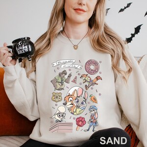 May include: Sand-colored sweatshirt featuring a colorful graphic of characters and elements from a popular animated movie. The design includes a donut, a police badge, and the text "You can be whatever you wish to be."