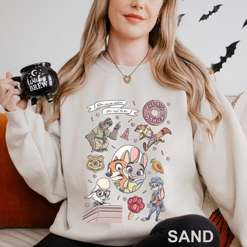 May include: Sand-colored sweatshirt featuring a colorful graphic of characters and elements from a popular animated movie. The design includes a donut, a police badge, and the text "You can be whatever you wish to be."
