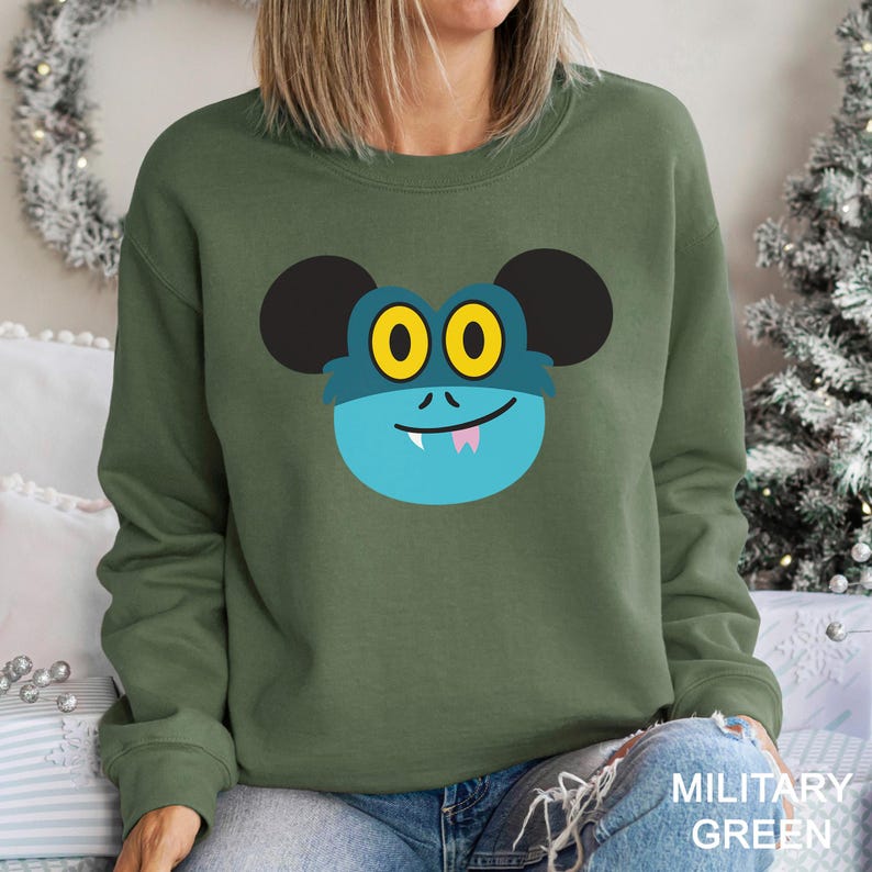 May include: Olive green sweatshirt featuring a cartoon monster face with large yellow eyes, a blue face, and black ears. The monster has a small pink tongue and a smiling mouth. The text "MILITARY GREEN" is printed at the bottom.