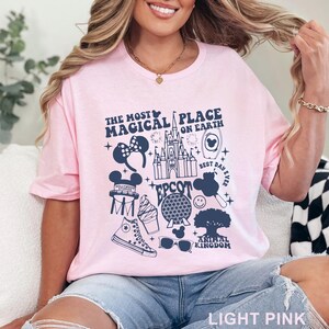 May include: Light pink t-shirt with a navy blue graphic featuring the text "THE MOST MAGICAL PLACE ON EARTH" and various Disney-themed icons. The design includes a castle, Mickey Mouse ears, and other park-related imagery.
