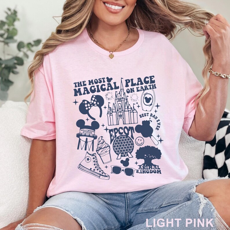 May include: Light pink t-shirt with a navy blue graphic featuring the text "THE MOST MAGICAL PLACE ON EARTH" and various Disney-themed icons. The design includes a castle, Mickey Mouse ears, and other park-related imagery.