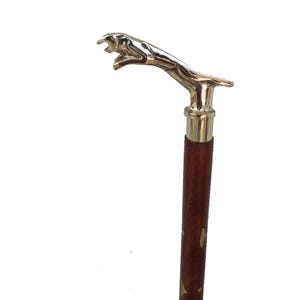 May include: A walking stick featuring a brass jaguar head handle. The shaft is made of dark brown wood with gold-coloured accents. A decorative and stylish accessory.