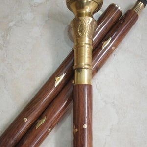 May include: A wooden walking cane with a brass handle and accents. The cane is dark brown with decorative brass inlays. The handle is ornate with a detailed design. The cane is partially disassembled, showing the individual sections.
