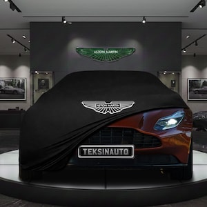 Può includere: Un telo nero per auto parzialmente drappeggiato su un'auto sportiva rossa. Il logo Aston Martin è visibile sul telo e sopra. La targa dice TEKSINAUTO.