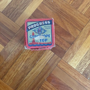 May include: A vintage "Progress Gyroscope Top" box, British made, with a red and blue color scheme. The box features the text "Progress" and "Gyroscope Top" in white and blue, respectively. The box is on a wooden surface.