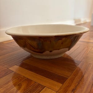 May include: A ceramic bowl with a cream-colored interior and a brown, textured exterior. The bowl has a wide, shallow shape and sits on a flat base. The exterior has a glazed appearance, with darker brown streaks.