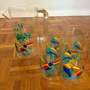 Puede incluir: Una jarra de plástico transparente con tapa y asa, y cuatro vasos a juego. Cada pieza está decorada con coloridos diseños de tucanes y hojas en rojo, amarillo, azul y verde.
