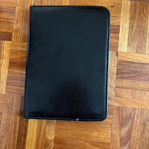 May include: A black, rectangular wallet with a textured, snake-skin-like pattern. The wallet is closed and appears to be made of leather or a similar material. The edges are neatly stitched.