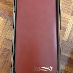 May include: A reddish-brown document holder with the word "Documents" in gold lettering. The holder is rectangular and is inside a cardboard box. The holder appears to be made of leather or a similar material.
