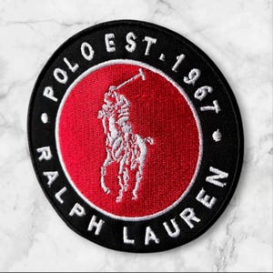 May include: A circular black patch with a red center featuring a white embroidered polo player on a horse. The patch is encircled by the words "POLO EST. 1967 RALPH LAUREN" in white lettering.