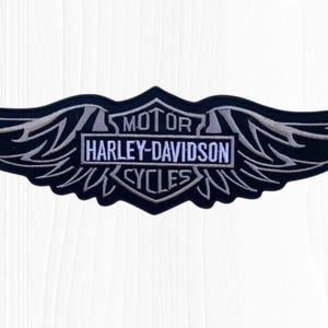May include: Black and silver Harley-Davidson patch in a wing design. The patch features the words "MOTOR HARLEY-DAVIDSON CYCLES" in a rectangular frame. The wings are detailed with silver stitching.