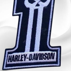 May include: A black and white Harley-Davidson patch in the shape of the number one. The number one features a skull design and the Harley-Davidson logo at the bottom. The patch has a white border.