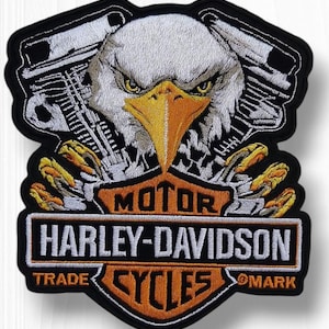 May include: Embroidered patch featuring a detailed eagle head with yellow eyes and beak, set against a black background. The Harley-Davidson logo is prominently displayed in orange and white, with the words "MOTOR HARLEY-DAVIDSON CYCLES".