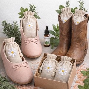 May include: A display of natural shoe sachets with a daisy design, presented in shoes and a box. The sachets are in ballet flats and cowboy boots. A bottle of peppermint essential oil is also present. The text reads "NATURAL SHOE SACHETS".