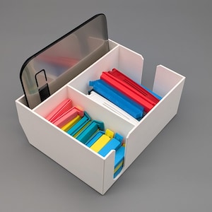 Könnte beinhalten: Ein weißer, rechteckiger Organizer mit mehreren Fächern. Ein Fach enthält ein Smartphone, andere enthalten farbige Plastiktütenclips in Rot, Blau, Gelb und Rosa. Der Organizer steht auf einer grauen Oberfläche.