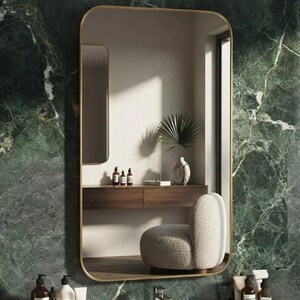 Puede incluir: Un espejo de pared rectangular con esquinas redondeadas y un marco dorado fino. El espejo refleja una habitación que contiene un mueble de madera flotante, una pequeña palmera en maceta y un sillón texturizado color crema sobre suelo de madera.