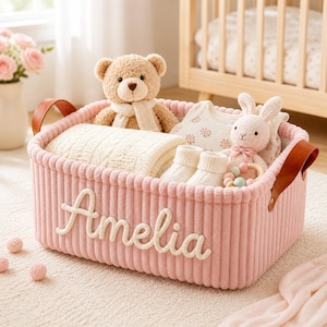Può includere: Un cesto portaoggetti in velluto a coste rosa con il nome "Amelia" in corsivo bianco. Il cesto contiene un orsacchiotto, una coperta, vestiti per bambini e un coniglietto di peluche. Il cesto ha manici in pelle marrone.