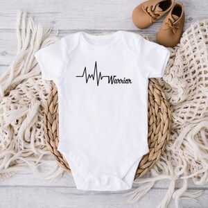 May include: White baby onesie with a black heartbeat line design and the word "Warrior". The onesie is short-sleeved and is placed on a woven basket with a cream-colored blanket and a pair of brown baby shoes.
