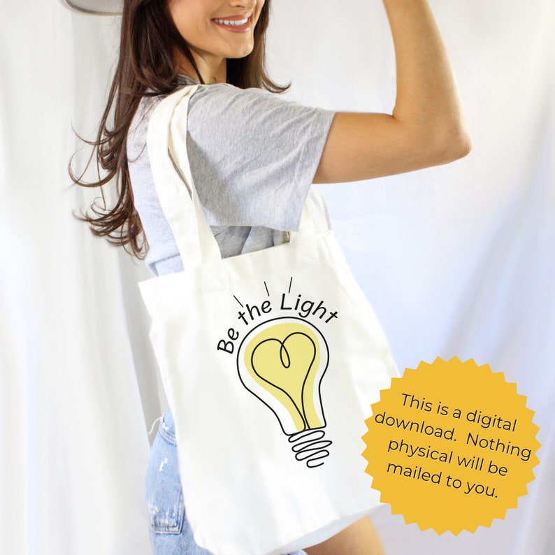 May include: A white tote bag with a light bulb graphic and the text "Be the Light". The light bulb is yellow with a heart shape in the center. The tote bag is being held by a person.