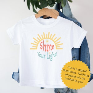 K&ouml;nnte beinhalten: Wei&szlig;es T-Shirt mit einem gelben Sonnen-Motiv und dem Text "Shine Your Light!" in bunten Buchstaben.