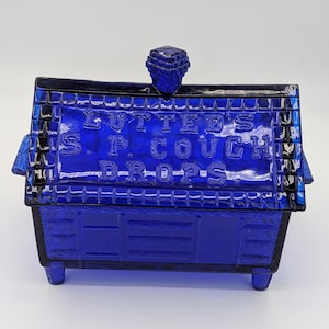 Puede incluir: Un recipiente de vidrio azul cobalto con forma de casa. El recipiente tiene una tapa con un pomo y las palabras "LUTTE'S S.P. COUGH DROPS" en relieve en la parte delantera. El recipiente se apoya sobre cuatro patas pequeñas.