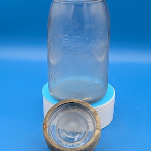 Könnte beinhalten: Ein klares, antikes Mason-Glas mit dem Wort "Mason's" auf der Vorderseite. Das Glas ist zylindrisch mit einer weiten Öffnung und einem separaten Deckel. Der Deckel ist aus Metall und trägt die Aufschrift "Clyde Glass Works".