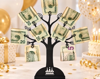 May include: A black metal tree-shaped money holder with a flat base featuring a white birthday cake icon. The branches have small clips holding several United States twenty-dollar bills and one five-dollar bill in a display arrangement.