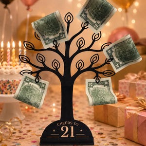 May include: A black metal tree-shaped money holder with a base engraved with the text CHEERS TO 21. The branches feature wire loops designed to hold paper currency, with four two-dollar bills currently attached to the display structure.