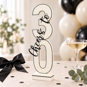 May include: A vertical cream-colored wooden sign featuring the number 30 with a black outline. The black cursive text "Cheers to" is layered over the number 30. The sign is mounted on a rectangular cream-colored base with a thin black line detail.