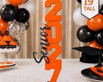 May include: A 19-inch tall orange wooden sign featuring the stacked numbers 2027 and the word Senior in black script lettering, mounted on a black rectangular base. The sign is displayed upright on a flat surface.