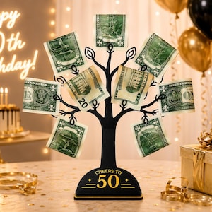 May include: A black metal money tree stand with a base featuring the text Cheers to 50. The tree has multiple branches designed to hold paper currency, with several US dollar bills clipped to the ends of the branches in a circular arrangement.