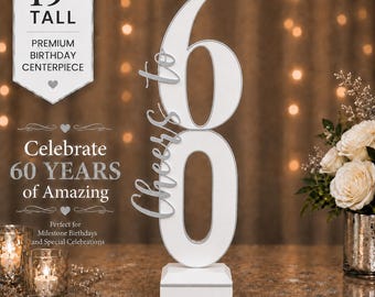 May include: A 19-inch tall white birthday centerpiece featuring the number 60 with the script text Cheers to attached to the side. The centerpiece stands on a square white base and includes silver-toned trim around the edges of the numbers.