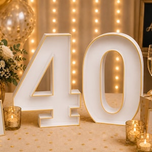 May include: A large white number 40 marquee sign with gold trim, displayed on a table alongside a bouquet of white roses in a gold vase, two champagne flutes, gift boxes, tea light candles, and two small signs reading Cheers to 40 wonderful years.