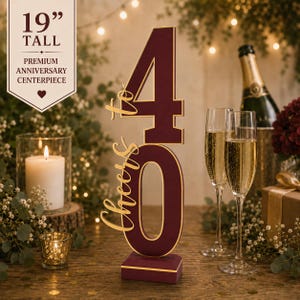 May include: A 19-inch tall burgundy and gold anniversary centerpiece featuring the number 40 and the script text Cheers to. The freestanding decoration has a rectangular base and a bold, block-style numeral design with a metallic gold outline.