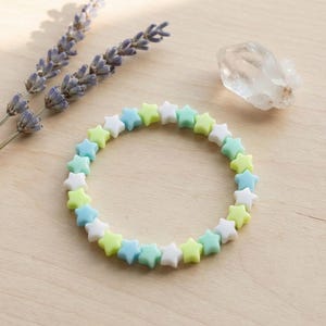 Könnte beinhalten: Ein Armband aus sternförmigen Perlen in Weiß, Hellblau und Gelbgrün. Das Armband ist kreisförmig und liegt auf einer hellen Holzoberfläche, neben Lavendelzweigen und einem klaren Kristall.