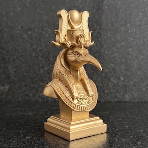 May include: A gold-colored bust of the Egyptian deity Thoth, depicted with an ibis head and a tall headdress featuring a crescent moon and solar disk. The sculpture rests on a square base and displays intricate carved details on the collar.