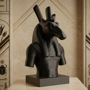 May include: A matte black bust of the Egyptian deity Anubis, depicted with a canine head, pointed ears, and a traditional nemes headdress. The sculpture features a detailed collar necklace and rests on a solid rectangular black base.