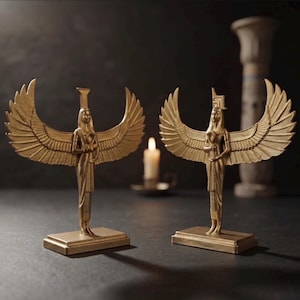 Könnte beinhalten: Zwei goldfarbene Statuen von geflügelten Figuren auf rechteckigen Sockeln. Jede Figur hat ausgebreitete Flügel, einen hohen Kopfschmuck und hält ein Ankh-Symbol in den Händen. Die Statuen sind mit detaillierten Texturen versehen.