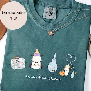May include: A blue spruce t-shirt featuring a graphic of four cartoon characters: a baby in an incubator wearing a witch hat, a baby bottle dressed as a vampire, a nasal aspirator, and a pill bottle holding a pumpkin bucket. The text reads nicu boo crew.