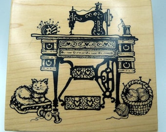 May include: A wooden stamp featuring a black illustration of a vintage sewing machine, a cat, a basket of yarn, and sewing notions. The design includes decorative hearts and spools of thread, perfect for crafting projects.