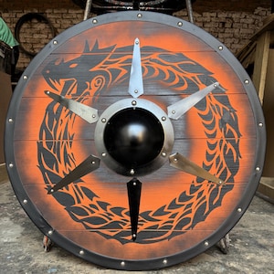 May include: A circular wooden shield with a black border and a central black boss. The shield has an orange background with a black dragon design. Metal accents and rivets complete the design, creating a striking Viking aesthetic.