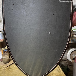 Peut inclure: Un bouclier noir avec une surface texturée et une bordure cuivrée. Le bouclier a un sommet arrondi et un fond pointu. Plusieurs rivets sont visibles sur la surface du bouclier. Il s'agit probablement d'un objet décoratif ou historique.