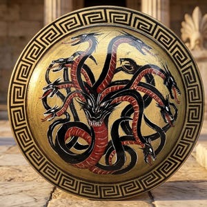 Puede incluir: Un escudo circular con una superficie dorada y una ilustración central de una hidra de varias cabezas en negro y rojo. El borde está decorado con un patrón geométrico griego en negro sobre un fondo dorado, creando un fuerte contraste.