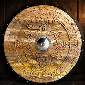 Peut inclure: Un bouclier rond en bois avec des sculptures complexes et un bossage central en métal. Le bouclier présente des motifs en relief détaillés, notamment des entrelacs et des figures animales stylisées, avec une bordure sombre et des clous métalliques. Le bois a une teinte chaude et naturelle.