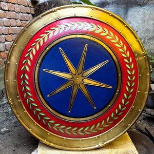 Op de afbeelding: Een rond schild met een blauw middenveld en een gouden achtpuntige ster. Het midden is omringd door een brede rode ring versierd met een groen lauwerkranspatroon. De buitenrand is een gouden metalen rand voorzien van kleine klinknagels.