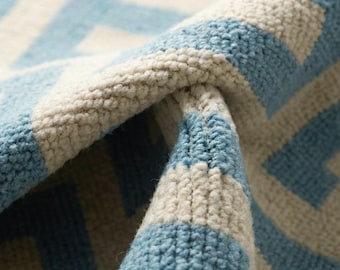 May include: Close-up of a woven rug with a geometric pattern. The rug features a repeating design of light blue and off-white shapes against a neutral background. The texture of the rug is visible, with a slight fold revealing the thickness of the material.